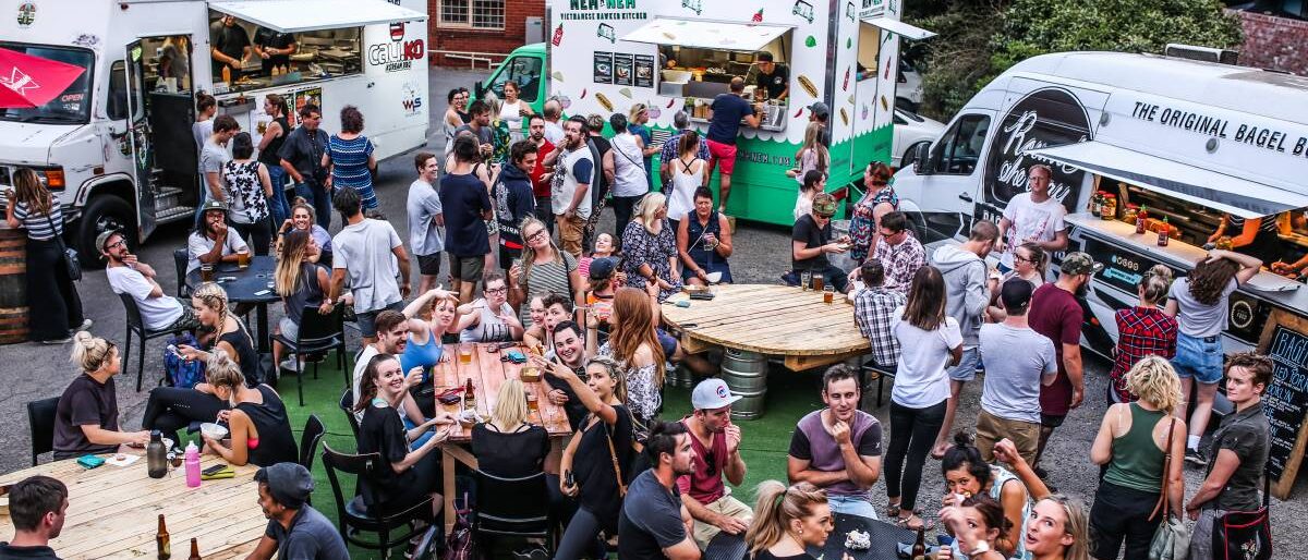 Four food trucks parked side by side in a yard, with a crowd of people queuing, drinking and eating at long communal tables between them.