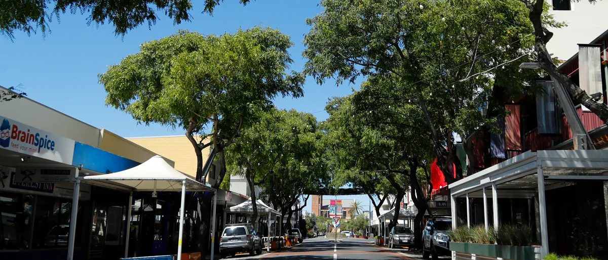 Victoria Street, Bunbury, looking down the centre of the road beneath a mature tree canopy, with awnings, festoon lighting and planter boxes along both sides and no people in sight.